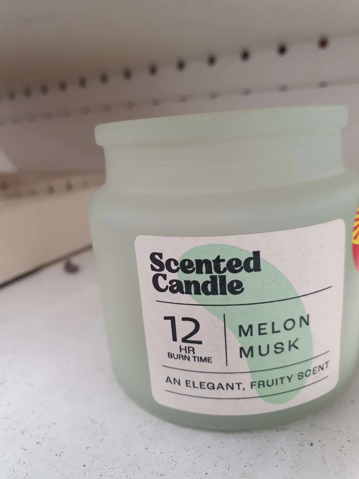 A close-up photo of a frosted glass jar candle on a store shelf, with a white label that reads 'Scented Candle', '12 HR BURN…