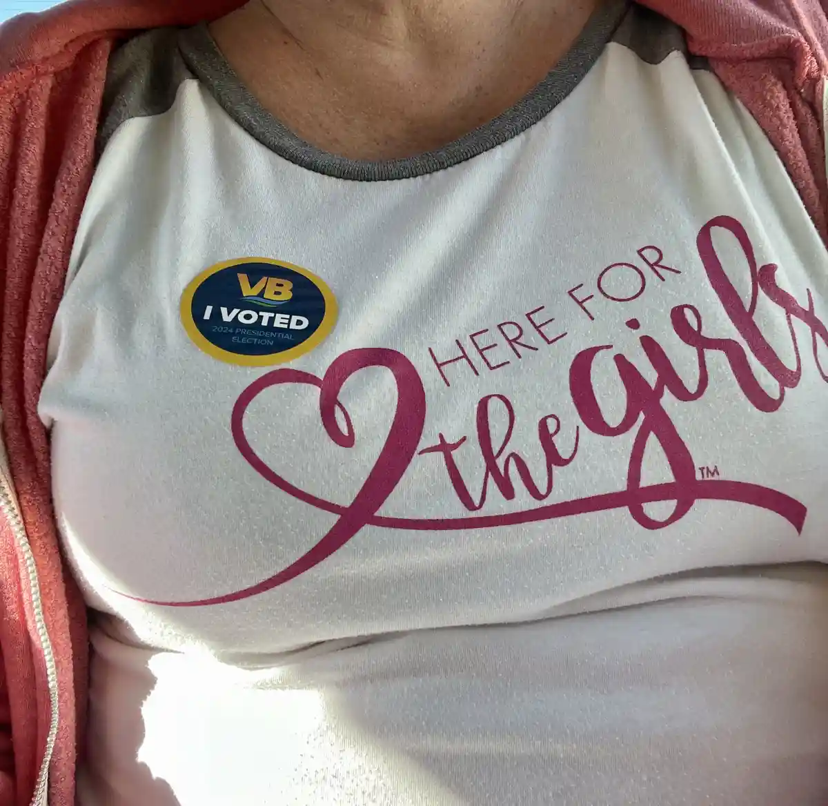 A close-up of a person wearing a white t-shirt with pink text reading 'HERE FOR the girls' and a heart symbol, with a yellow…