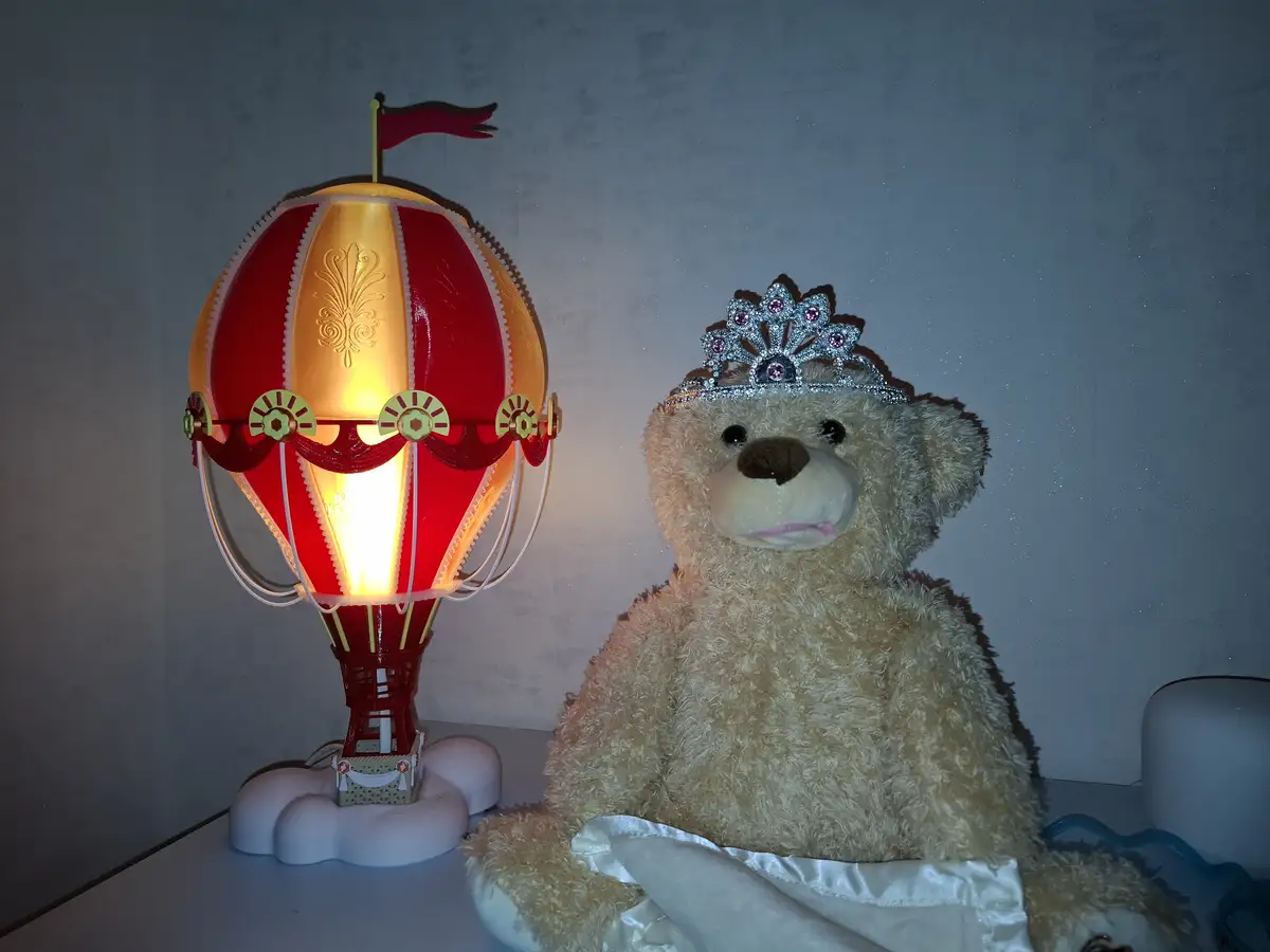 A beige stuffed teddy bear wearing a sparkly tiara and white satin skirt sits next to a glowing red and gold hot air…