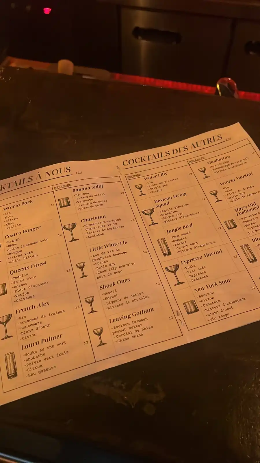 A dimly lit cocktail menu is displayed on a dark bar counter, featuring two sections: 'Cocktails à Nous' (created by the…