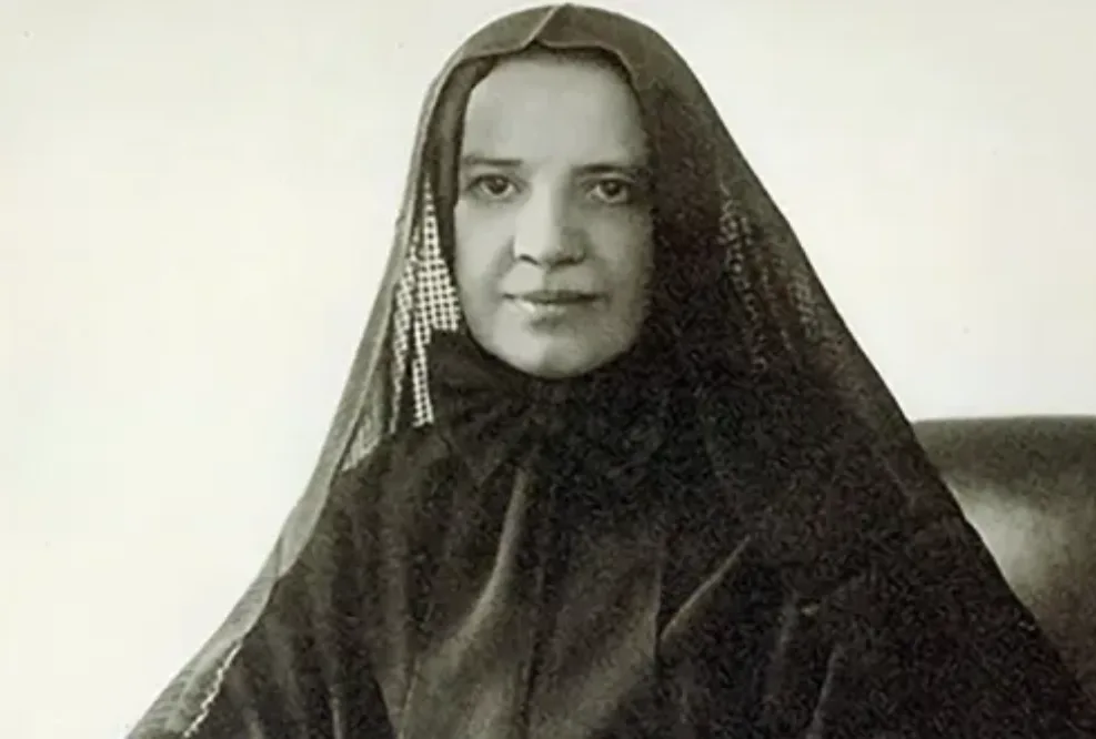 A vintage black-and-white portrait of a woman wearing a dark headscarf with a checkered pattern along the edge, seated…