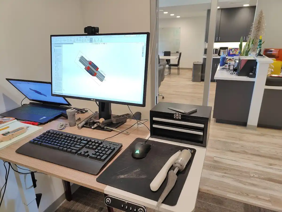 A desk setup with a large monitor displaying a 3D CAD model of RAM sticks, a laptop, keyboard, mouse, and a small black…