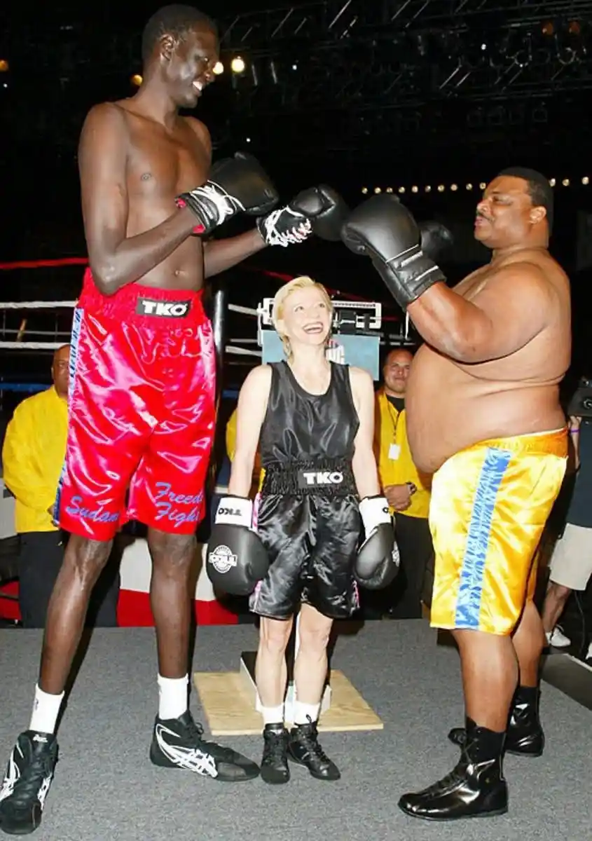 Three people in boxing gear stand in a ring: a tall Black man in red shorts, a short blonde woman in black shorts, and a…