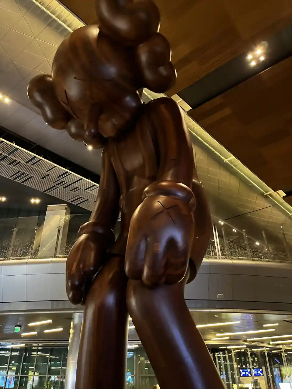 A large, glossy brown wooden sculpture of a stylized cartoon character with a sad face and X's for eyes, standing in a…