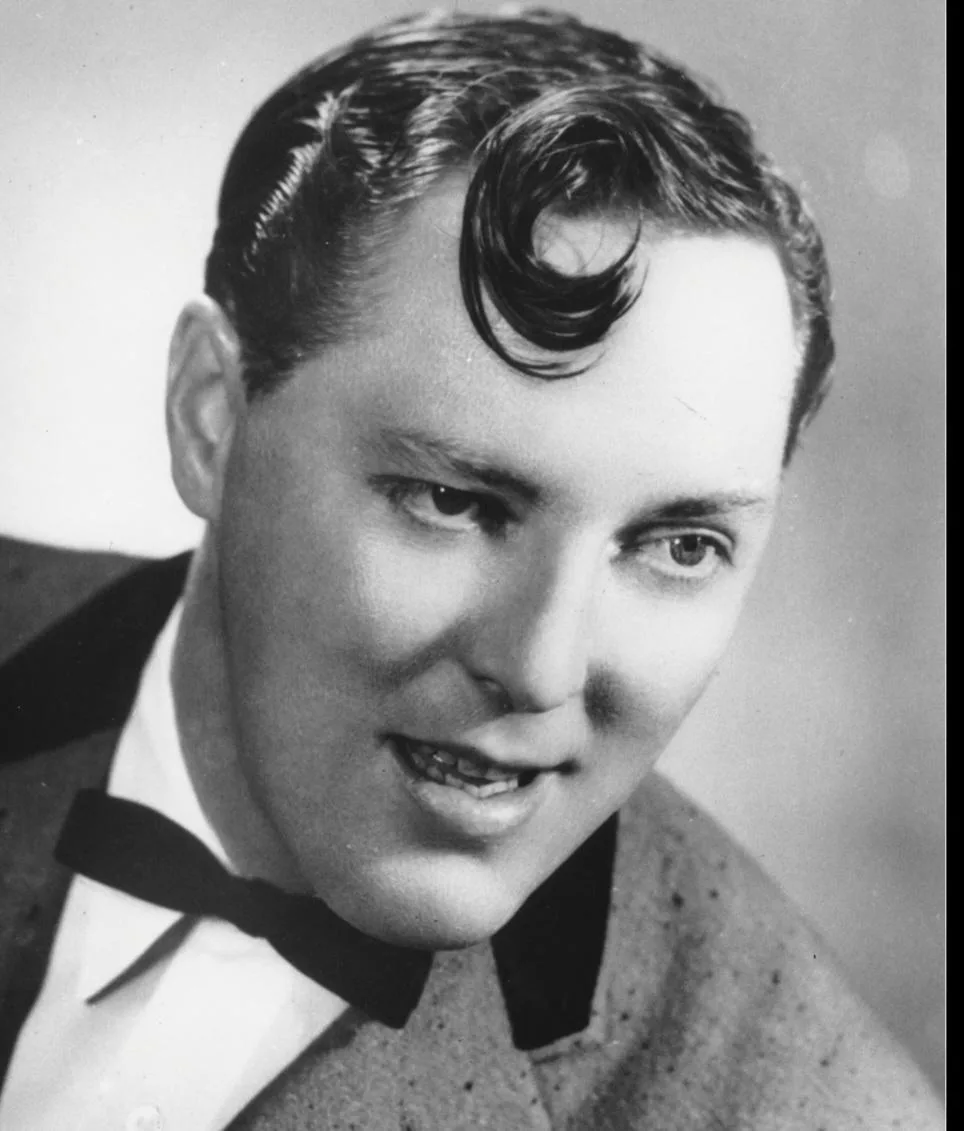 A black-and-white portrait of a man with a prominent, curled side part hairstyle, wearing a suit and bow tie, with the…