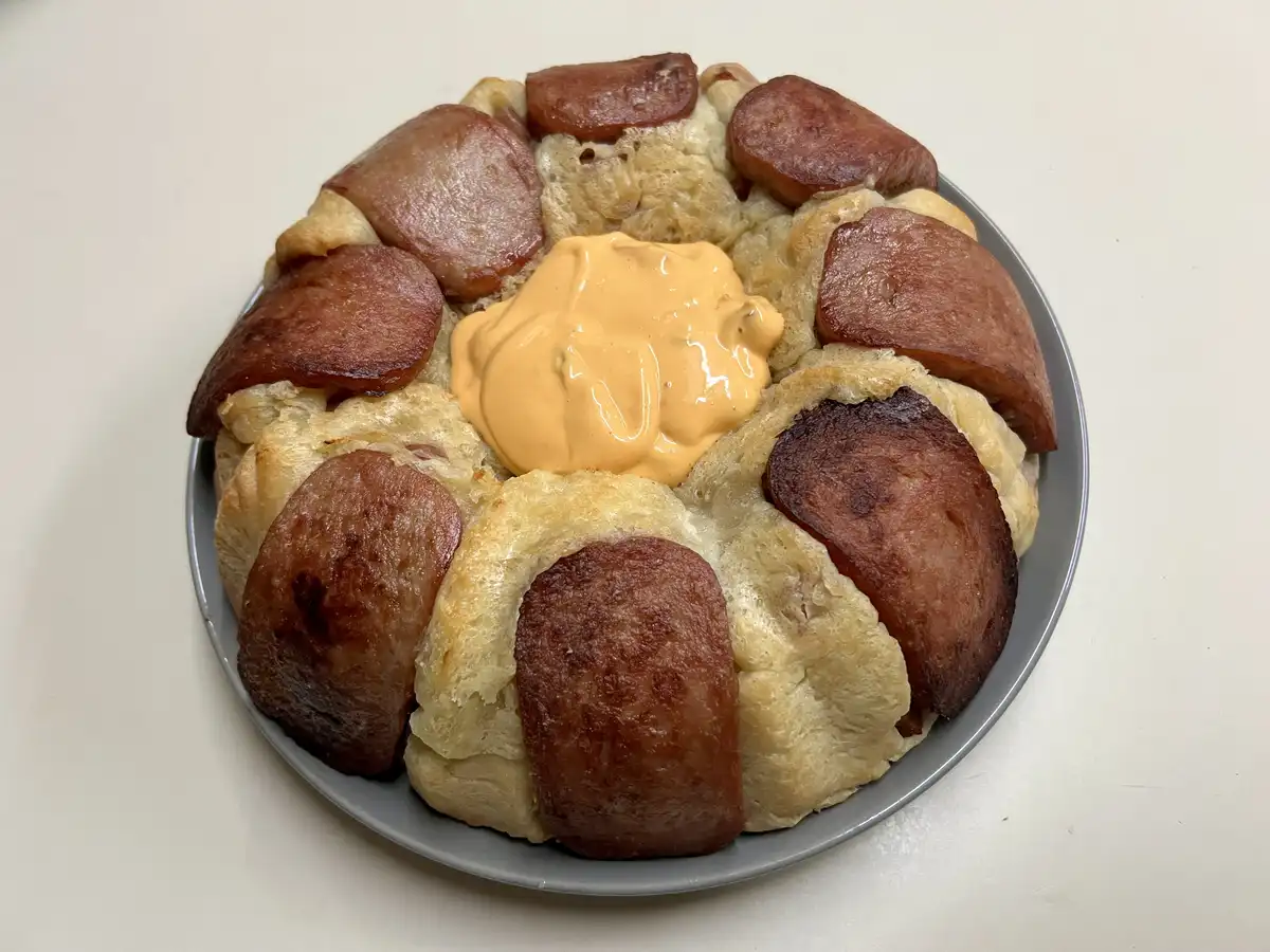 a round baked bread ring with golden-brown pastry segments, each topped with a slice of cooked sausage, arranged in a circle…