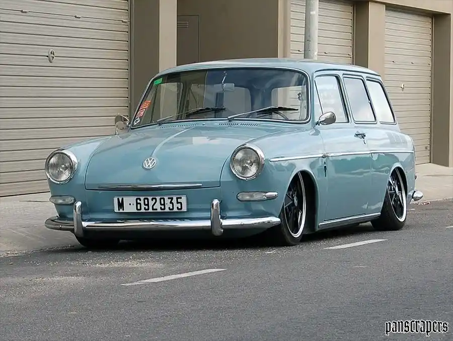 A light blue vintage Volkswagen Type 3 station wagon is parked on a street in front of beige garage doors. The car has been…