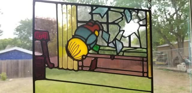 A stained glass window depicts a cartoonish figure with a yellow head and blue body, seemingly running or falling backward…