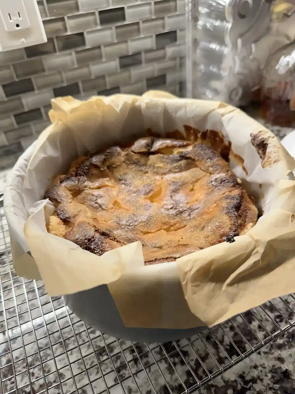 A close-up photo shows a round baked dessert in a greased pan lined with parchment paper, resting on a cooling rack. The…