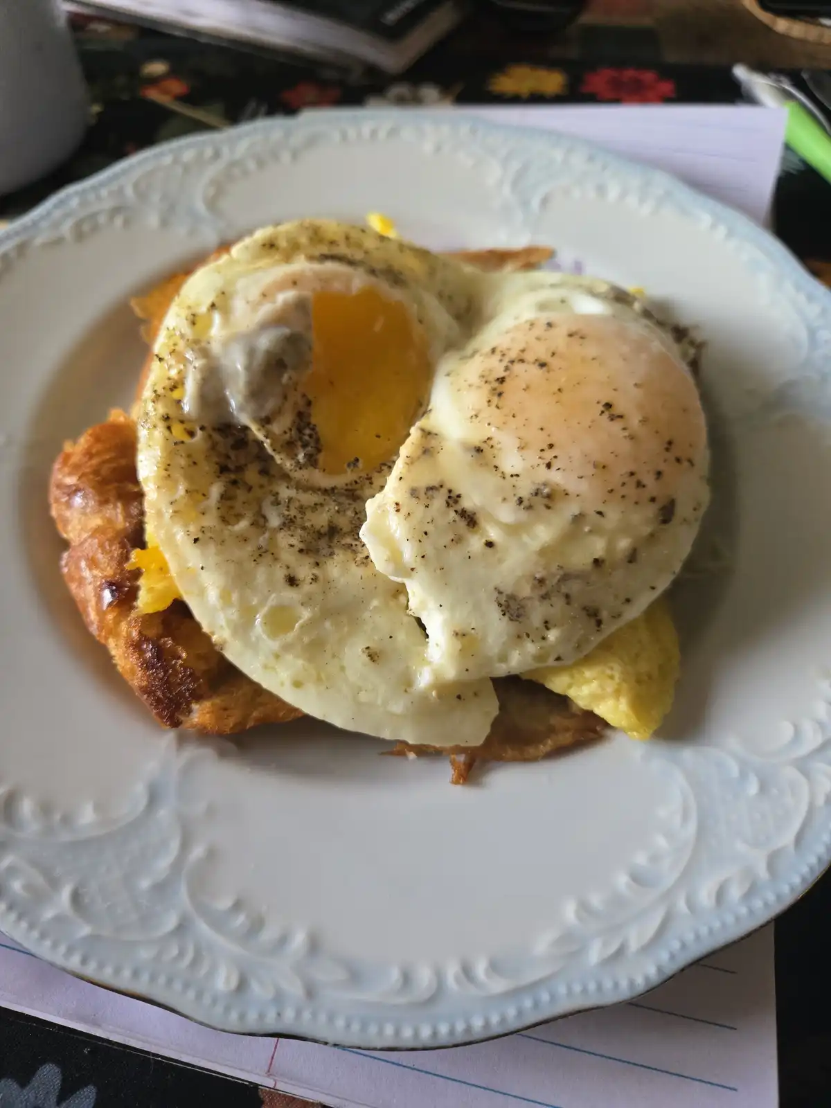 A close-up photo shows a breakfast dish on a white ornate plate: two fried eggs, one with a runny yolk, sit atop a croissant…