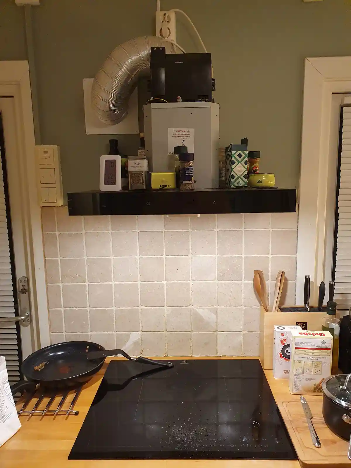 A kitchen scene shows a black induction cooktop with a dirty frying pan resting on a cooling rack to its left. Above the…