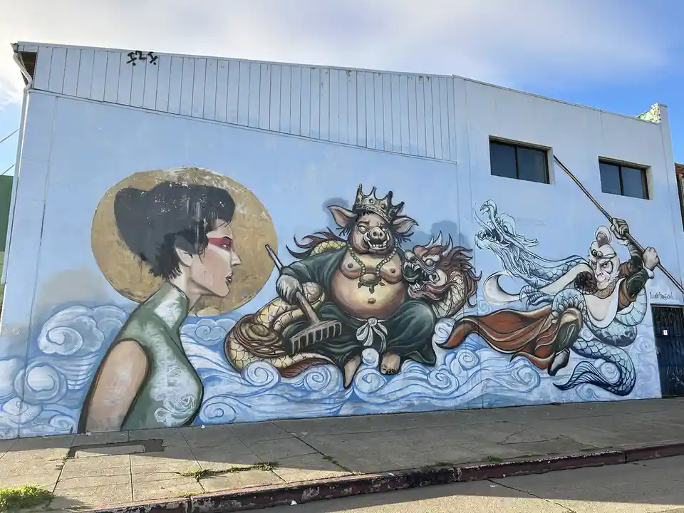A mural on a light blue building depicts three mythological figures: a woman in profile with a golden halo, a crowned…