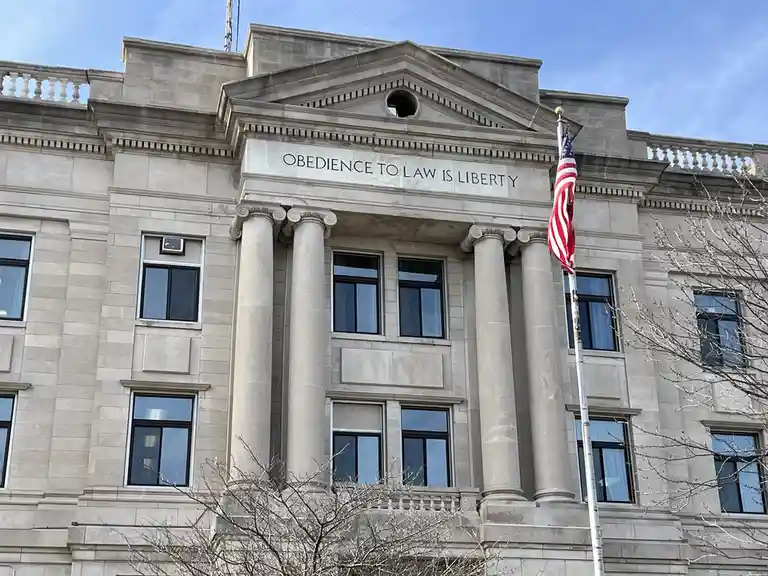 A photo of a classical stone courthouse facade with large columns and a pediment. Inscribed above the entrance is the phrase…