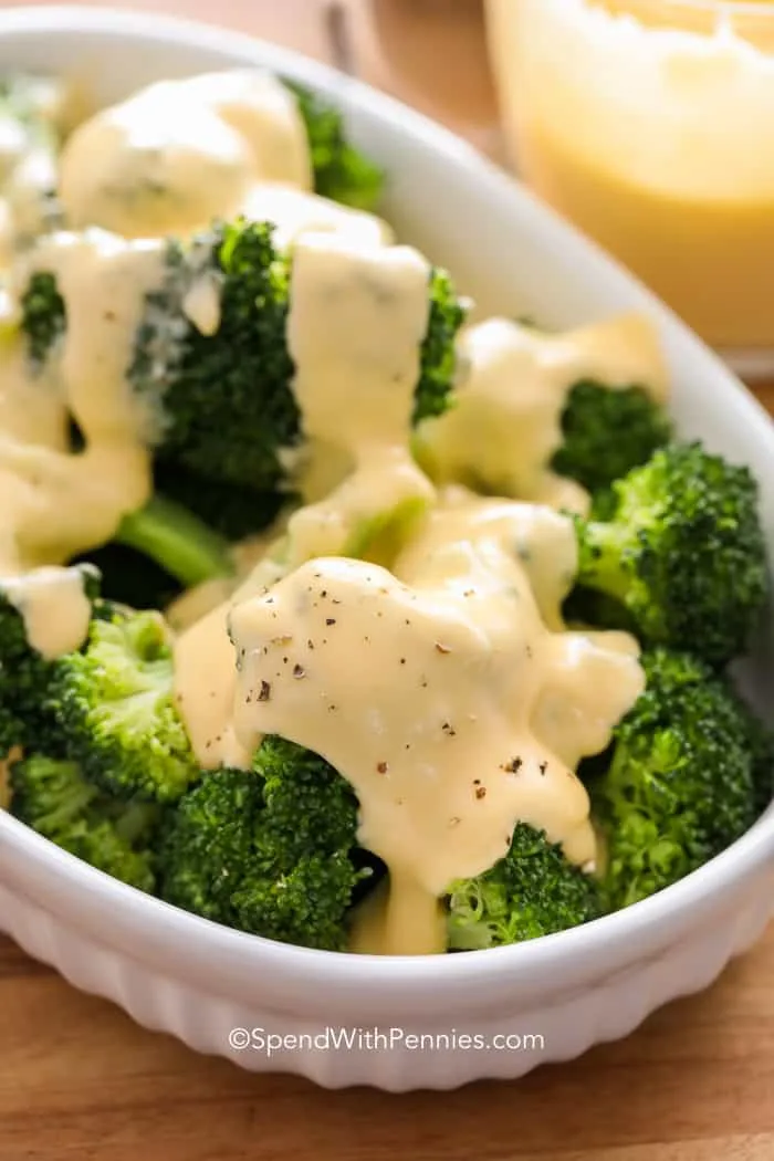 A close-up photo shows bright green broccoli florets in a white ceramic bowl, generously covered in a creamy, pale yellow…