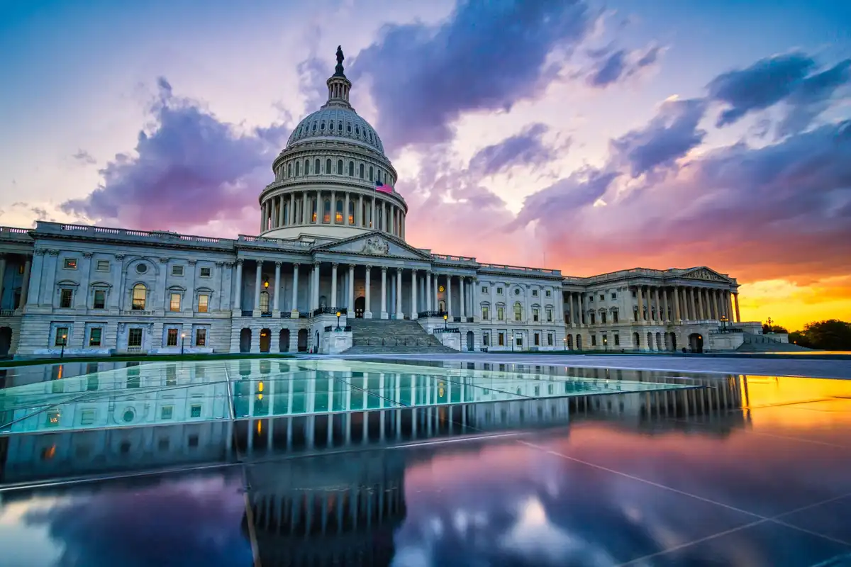 A stunning photograph of the U.S. Capitol Building at sunset, with vibrant purple and orange clouds reflected in a large…