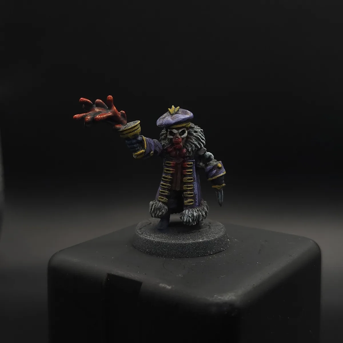 A painted miniature figurine of a skeletal clown with a purple hat, red nose, and elaborate coat stands on a grey base…