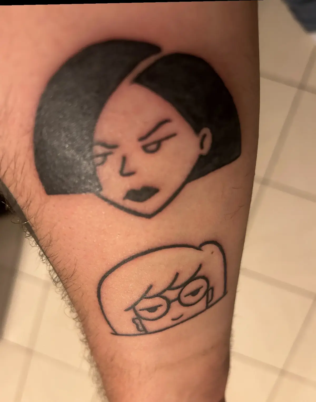 A close-up photo shows two tattoos on a person's leg: the top one is a black-and-gray stylized portrait of a woman with a…