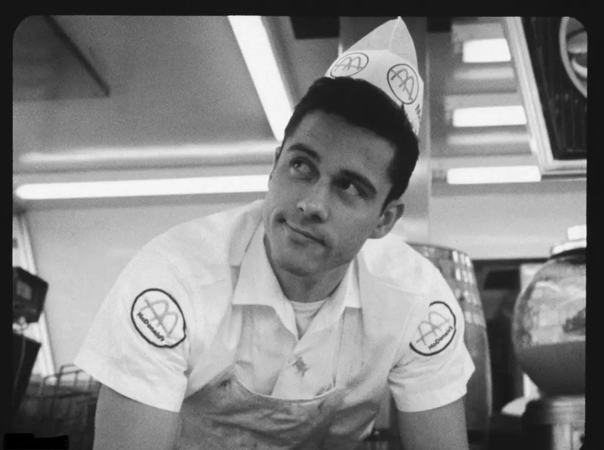 A black-and-white photo of a young man in a vintage McDonald's uniform, including a paper hat and logoed shirt, looking…