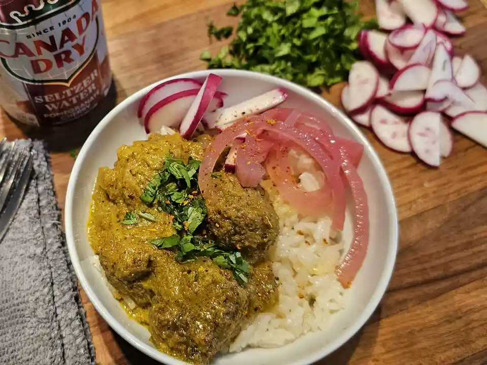 A white bowl of curry with meatballs, rice, pickled onions, and sliced radishes sits on a wooden table next to a can of…