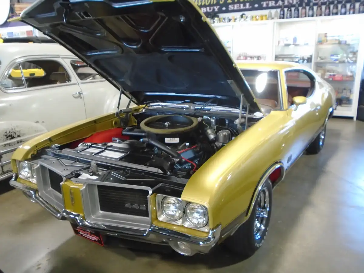 A yellow classic muscle car with its hood open, revealing a detailed engine bay, parked inside a garage with another vintage…