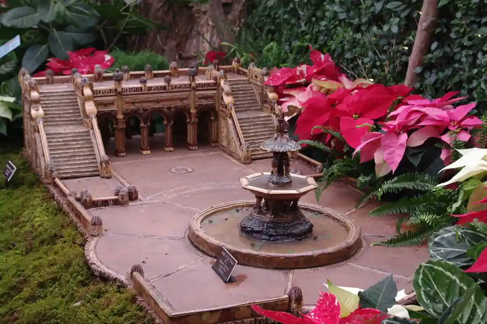 A miniature model of the Bethesda Fountain in Central Park, surrounded by lush greenery and vibrant red poinsettias, with a…