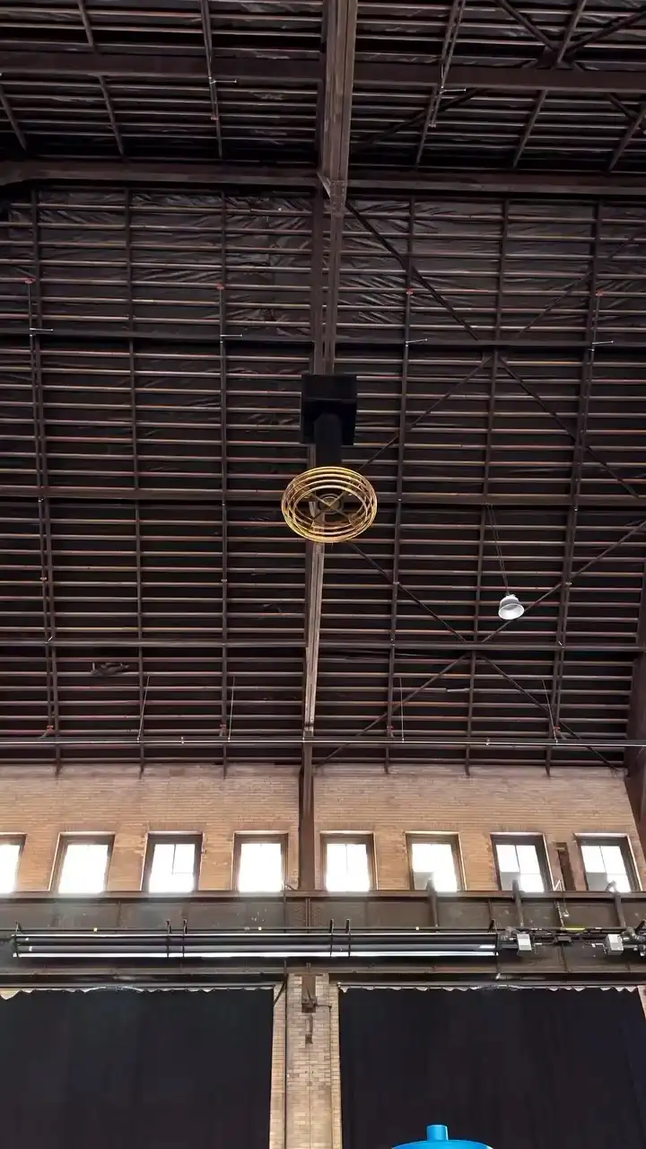 A low-angle shot looks up at a high, industrial-style ceiling with exposed metal beams and brick walls. Suspended from the…