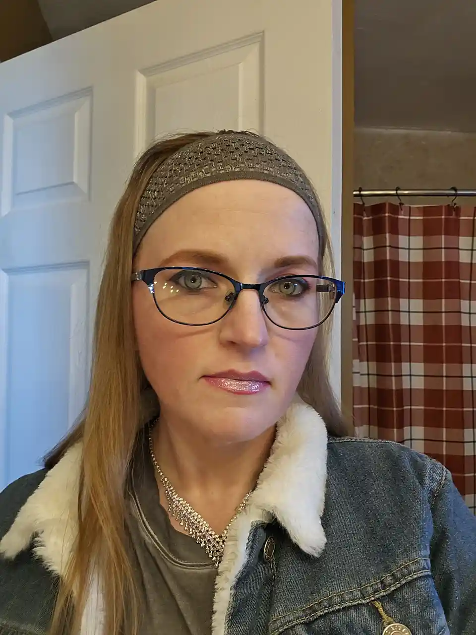A woman with glasses, a headband, and a denim jacket with a white fur collar stares directly at the camera with a neutral…