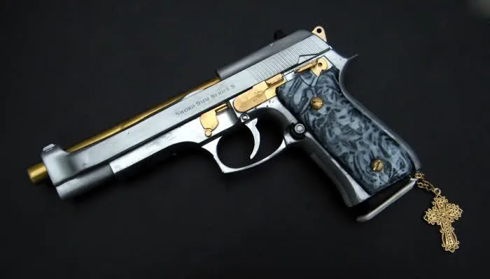 A silver and gold pistol with ornate blue grip and a gold cross pendant hanging from its trigger guard, set against a black…
