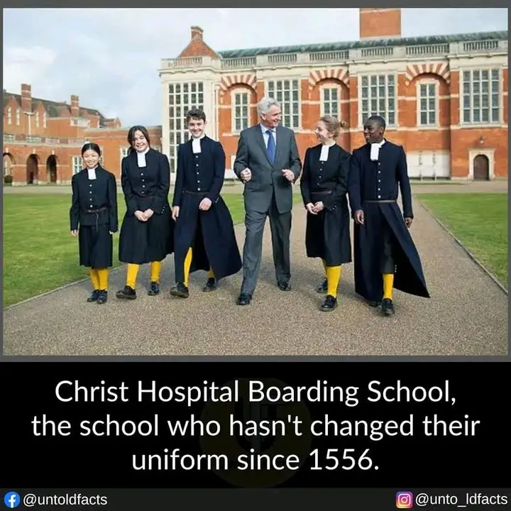 Six students in identical black academic uniforms with white collars and bright yellow socks walk alongside a man in a suit…