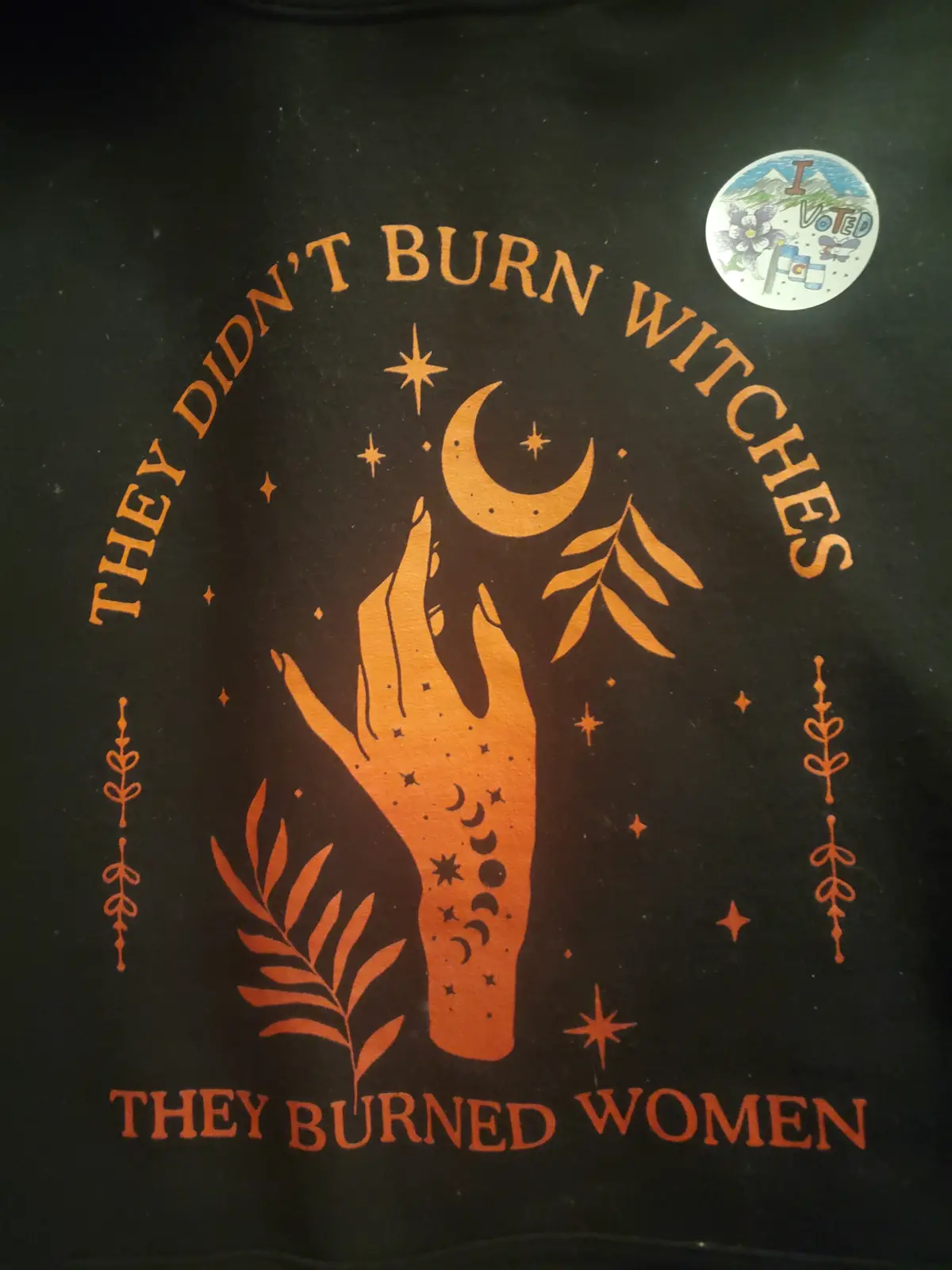 A black t-shirt features orange text and graphics, including the phrase 'THEY DIDN'T BURN WITCHES THEY BURNED WOMEN' arched…
