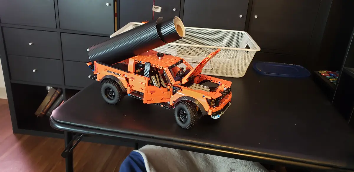 An orange LEGO Technic Ford Raptor truck model sits on a black desk with its hood open, and a large roll of black carbon…