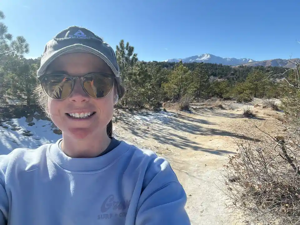 A person wearing a cap and sunglasses smiles at the camera while standing on a snowy trail with mountains in the background…