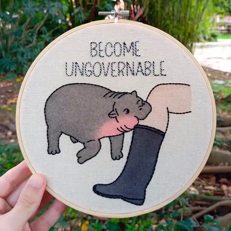 An embroidery hoop holds a stitched design of a gray hippopotamus affectionately nuzzling a human leg wearing a black boot…