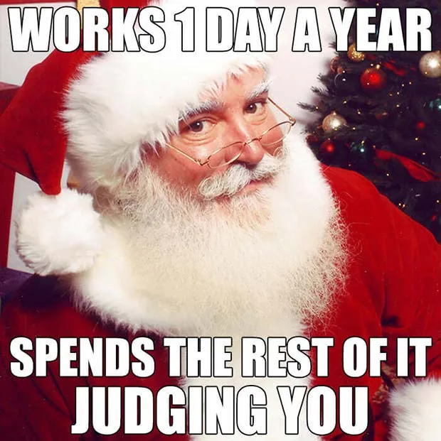 A man dressed as Santa Claus with glasses, a white beard, and a red suit sits in front of a decorated Christmas tree. The…