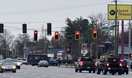 A street view with multiple traffic lights showing red, cars stopped at the intersection, and a yellow Planet Fitness sign…