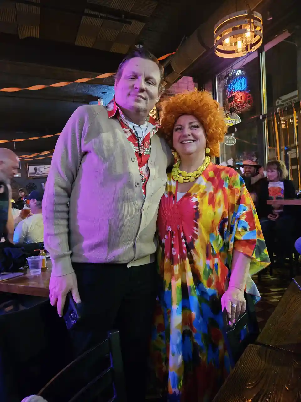 A man and a woman pose together in a dimly lit bar with colorful decor. The woman wears a vibrant, multi-colored tie-dye…