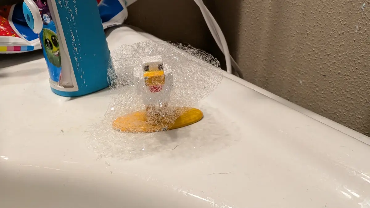 A small, blocky, yellow and white duck figurine stands on a white bathroom counter, completely encased in a large…