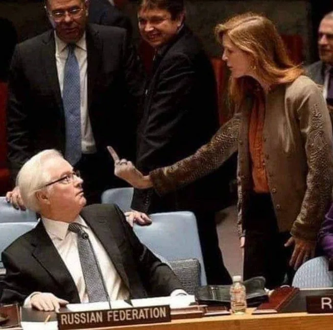 a woman in a beige cardigan and orange top stands over a man seated at a desk labeled 'RUSSIAN FEDERATION', pointing her…