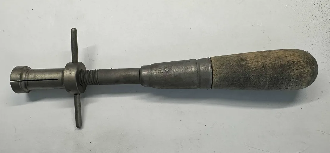 a close-up photo of a vintage metal tool with a worn wooden handle and a T-shaped adjustment knob, lying on a plain white…