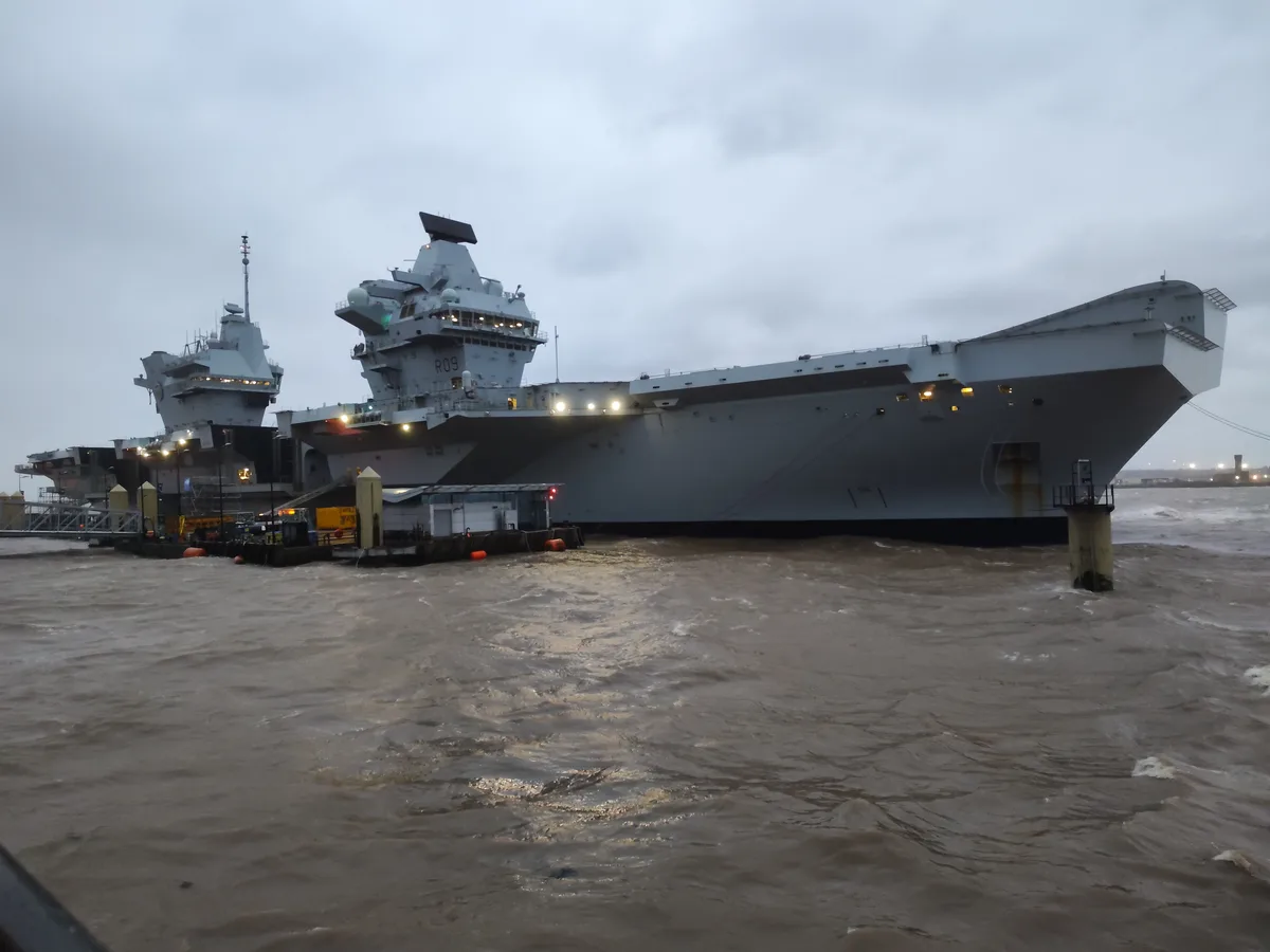 A large grey aircraft carrier, marked with 'R09', is docked at a pier under an overcast sky, with choppy, murky brown water…
