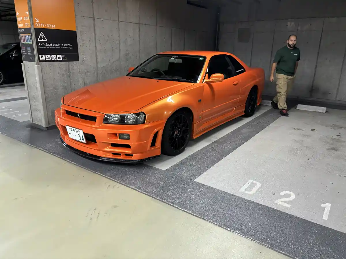 An orange Nissan Skyline R34 is parked in a concrete parking garage, its front bumper touching a stopper, with a man…