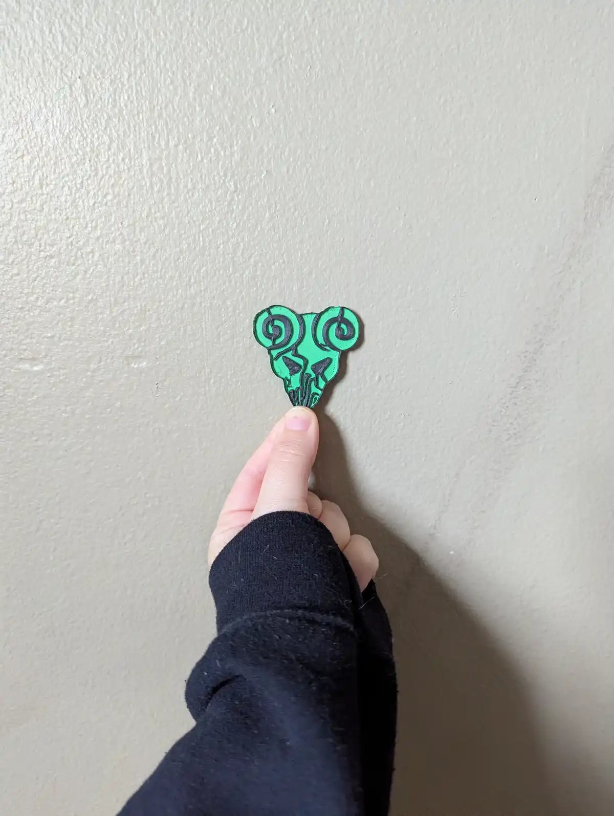 A hand holds a small green pin shaped like a stylized skull with spiral horns against a plain beige wall. The pin has black…