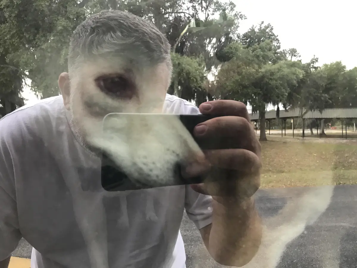 A man with short gray hair and a white t-shirt is taking a selfie through a fogged-up glass surface, holding a smartphone…