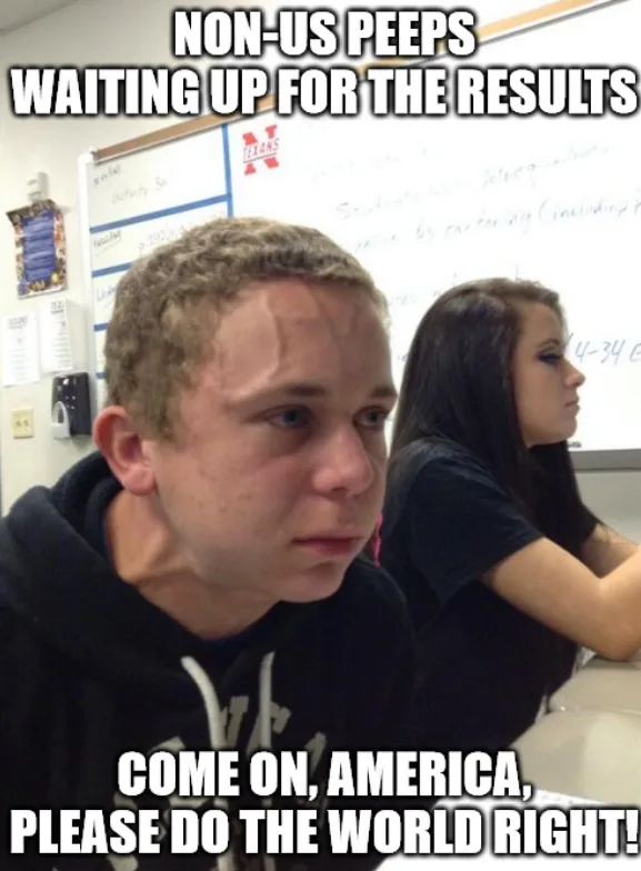 A young man with a bored, slightly annoyed expression sits in a classroom, looking off to the side. Behind him, a woman with…