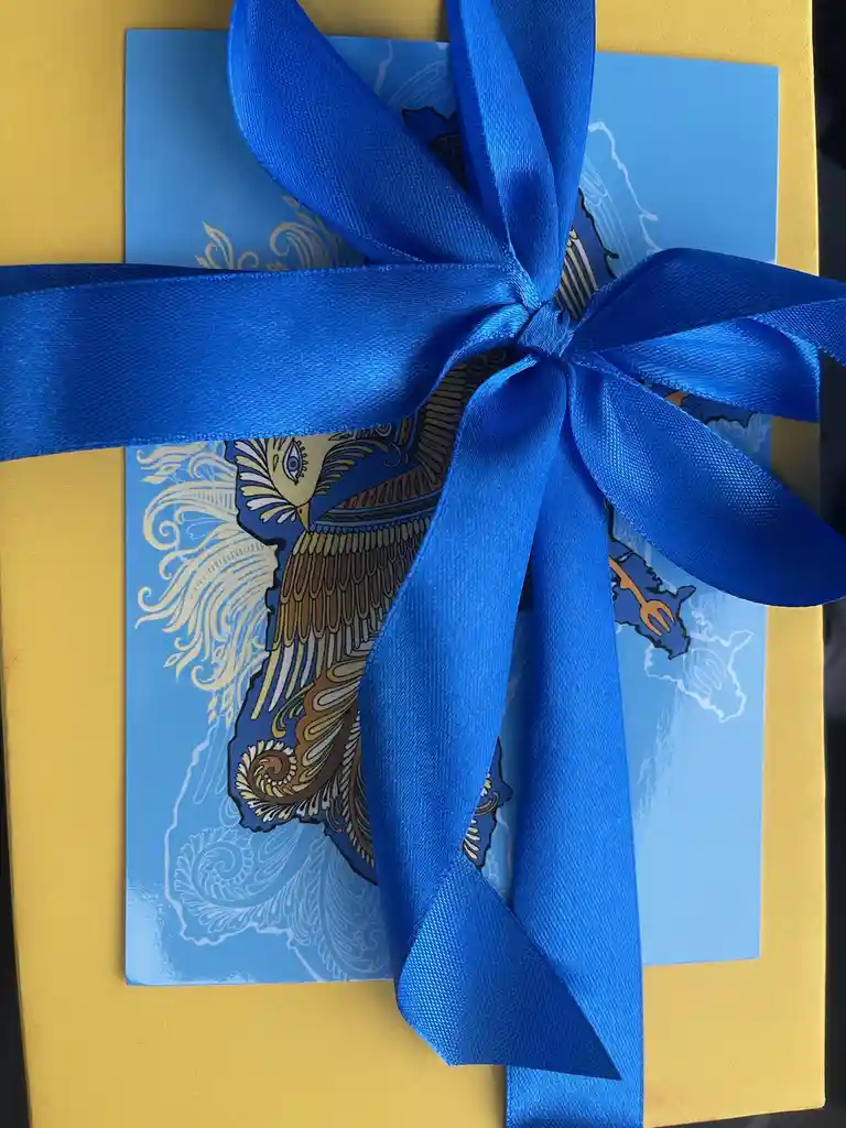 A gift wrapped in a yellow envelope with a blue card featuring an ornate bird illustration, tied with a large, shiny blue…