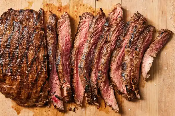 A top-down photo shows a grilled steak sliced into thin pieces on a wooden cutting board, revealing a pink, medium-rare…