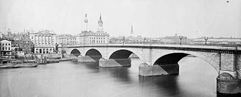 A black-and-white historical photograph shows a large stone arch bridge spanning a wide river, with European-style buildings…