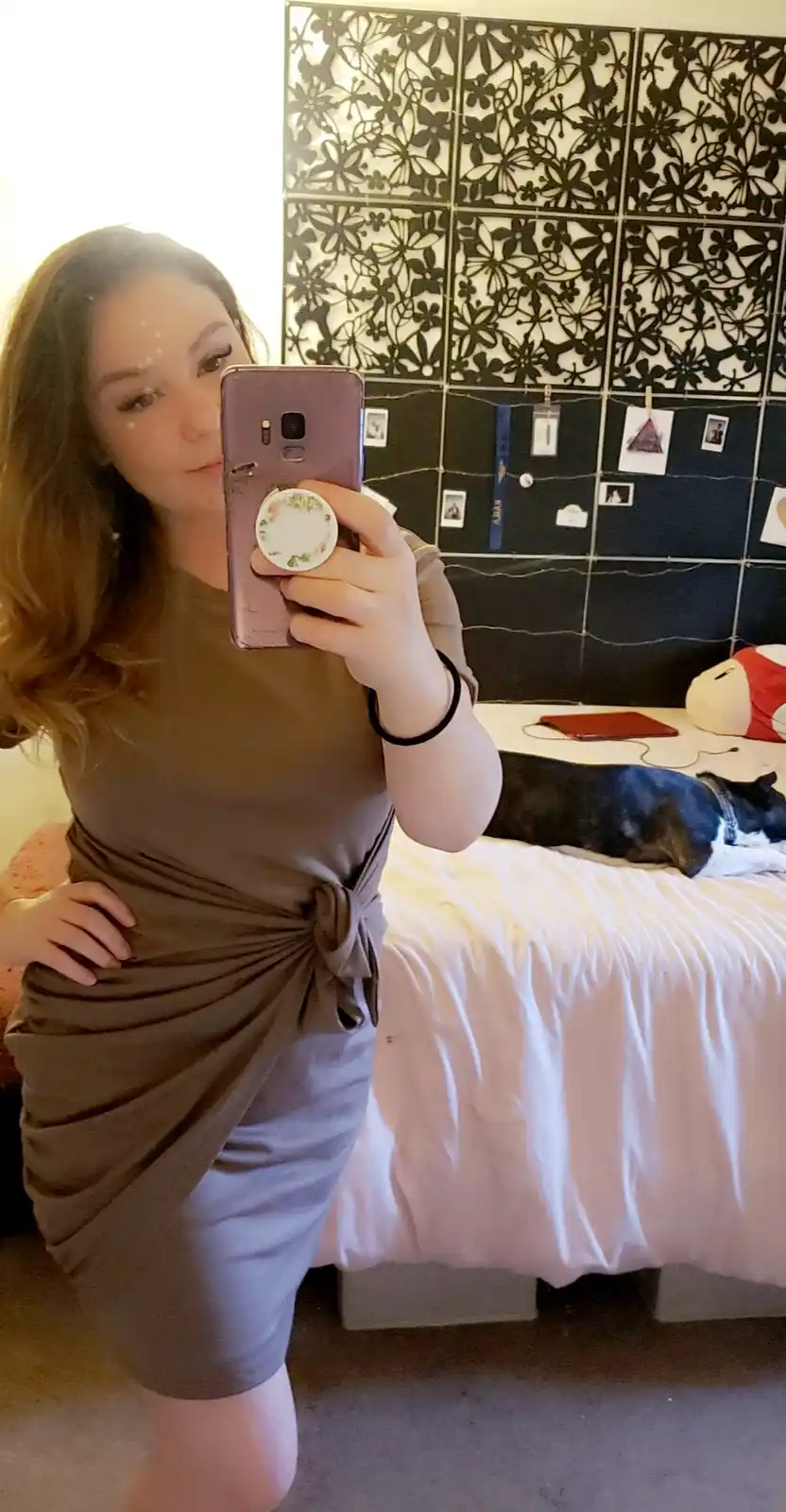 A woman takes a mirror selfie in a bedroom, wearing a brown dress tied at the waist, holding a phone with a floral pop…
