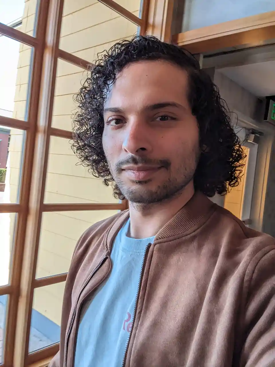 A selfie of a man with dark curly hair and a light beard, wearing a brown zip-up jacket over a light blue t-shirt, standing…