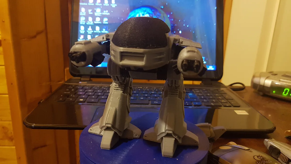 A gray 3D-printed robot figurine with a mesh head and two cannon-like arms stands on a blue base in front of an open laptop…